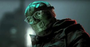 The Riddler character from The Batman movie, wearing a green mask, glasses, and a dark hooded jacket, set against a dimly lit urban background